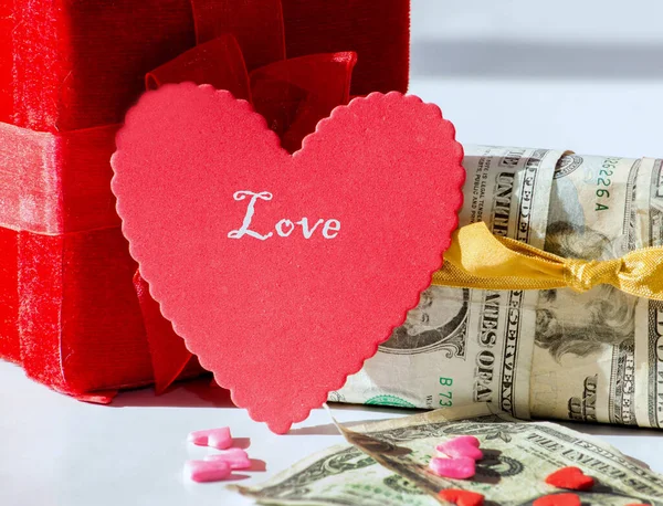 一颗红色的心和一个读取的礼品盒与另一个盒子裹在金钱 前景中的糖果 爱的代价 图库照片