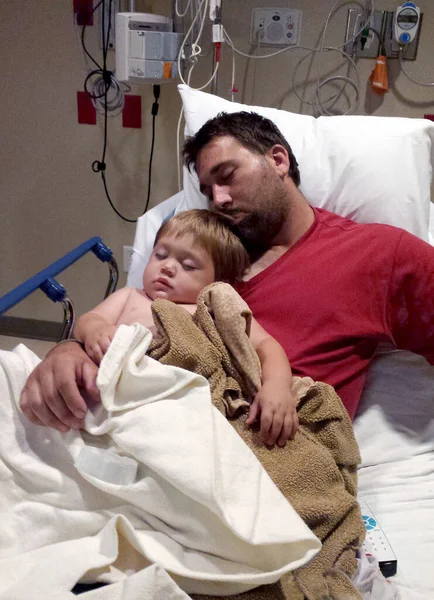 一个疲惫的爸爸在医院的病床上抱着他那蹒跚学步的儿子 图库图片