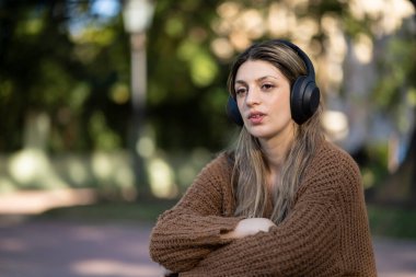 Parkta oturmuş düşünceli bir ifadeyle müzik dinleyen genç bir kadın.