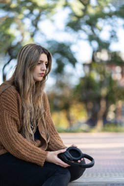 Parkta oturan genç bir kadın endişeli bir ifade ve kulaklık tutarken kaybolmuş bir bakış.