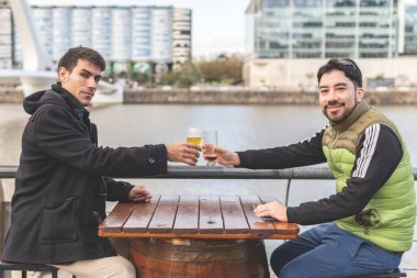 Latin turistler bir açık hava barında bira içiyor, kadeh kaldırıyor ve Puerto Madero, Buenos Aires, Arjantin 'de kameraya bakıyor.