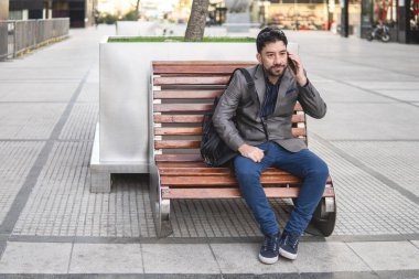 Latin iş adamı bankta oturmuş telefonda konuşuyor.