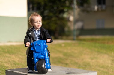 Bir yaşında sarışın ve mavi gözlü bebek parkta motosiklet sürüyor.