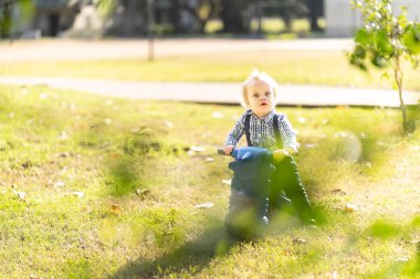 Bir yaşında sarışın ve mavi gözlü bebek parkta motosiklet sürüyor.