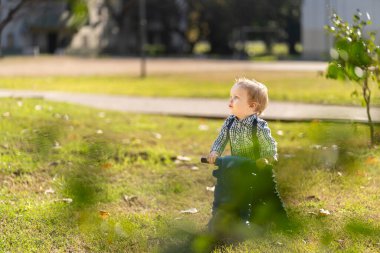 Bir yaşında sarışın ve mavi gözlü bebek parkta motosiklet sürüyor.