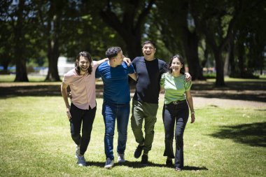 Parlak güneşin altındaki bir parkta kol kola yürüyen bir grup Latin arkadaş.