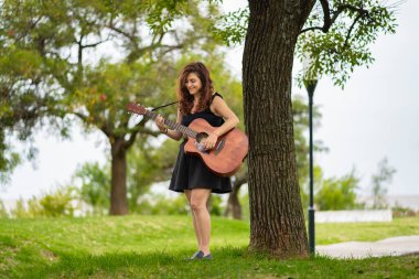 Genç bir kadın güzel bir parkta gitar çalarken gülümser.