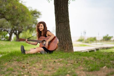 Genç Latin kız çimenlerde oturuyor gitar çalıyor ve şarkı söylüyor.