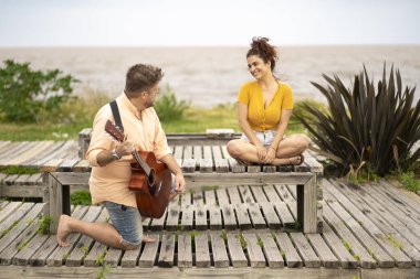 Sahilde dizlerinin üzerine çöküp kız arkadaşına gitar çalan bir adam.