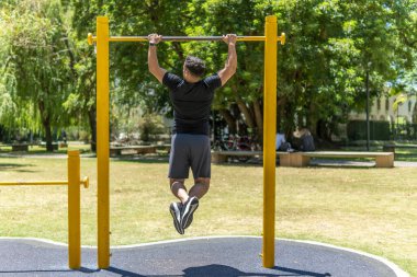 Genç bir adamın parktaki bir barikatın önünde barfiks çekişi.