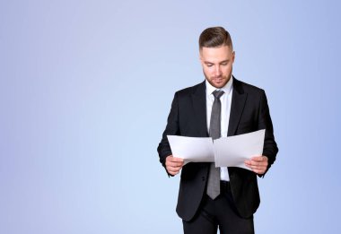 Resmî takım elbiseli yakışıklı işadamı arka plandaki boş soluk mavi duvarın yanında duran notları okuyor. Model kavramı, düşünen iş adamı, düşünülen adam, bilgi, belgeler