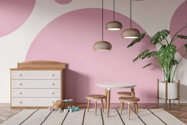 Front view on bright baby room with table with stools, sideboard, car toy, pink and white wall, oak wooden hardwood floor, carpet, plant. Concept of nursery in soft design, cozy space for newborn kid.