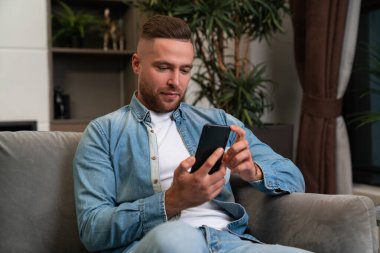 Concentrated young man using smartphone, sitting on sofa at home. Concept of social media and video call