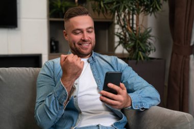 Glad handsome businessman wearing casual wear surfing net on smartphone sitting on sofa in background. Concept of smiling business person, pleased man, information, internet communication