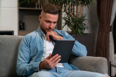 Serious handsome businessman wearing casual wear surfing net on tablet device sitting on sofa in background. Concept of considered business person, pondering man, information, internet communication