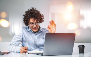Businessman wearing formal wear is having online video conference call via laptop sitting at office workplace in background, taking notes. Concept of distant working process, gadgets, communication