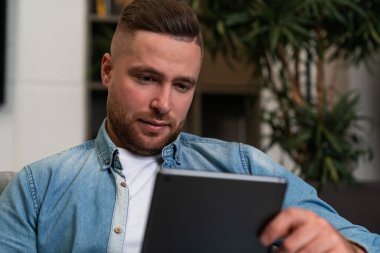 Serious man portrait looking at tablet in hand, denim shirt sitting on sofa. Concept of social media and online connection