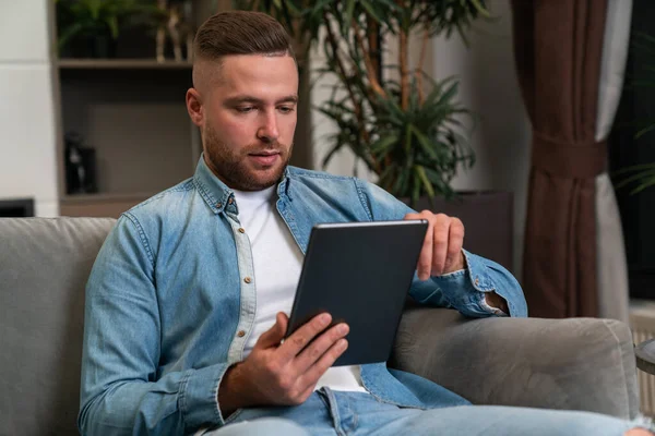 Serious man sitting on sofa at home with tablet in hand, busy working online. Concept of reading books, social media and video call