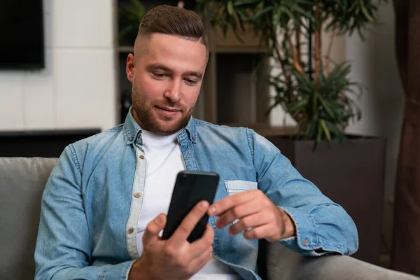 Dreaming handsome businessman wearing casual wear surfing net on smartphone sitting on sofa in background. Concept of information, internet communication, online meeting with friends