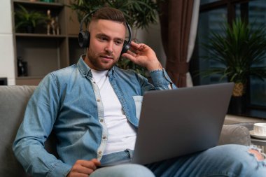 Thinking businessman in headphones looking at laptop, sitting on sofa at home. Concept of video conference and business meeting