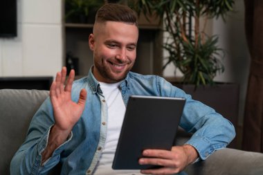 Happy young man waving hand and looking at tablet, online video conference and communication. Concept of distance work and mobile app