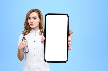 Young doctor woman show smartphone, blue background. Concept of telemedicine and consultation. Mock up copy space display