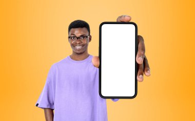 Young black man in eyeglasses smiling, showing phone with mock up copy space screen, yellow background. Concept of mobile app and social media