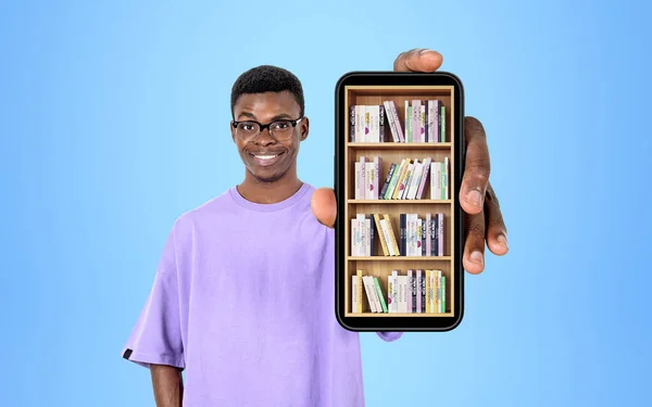 African American businessman wearing casual wear is showing smartphone case with digital library. Bookshelf with various books. Concept of e-learning and online education. Blue wall in background