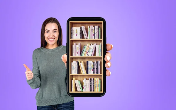 Young attractive businesswoman wearing casual wear is showing smartphone case with digital library. Bookshelf with various books. Concept of e-learning and online education. Purple wall in background