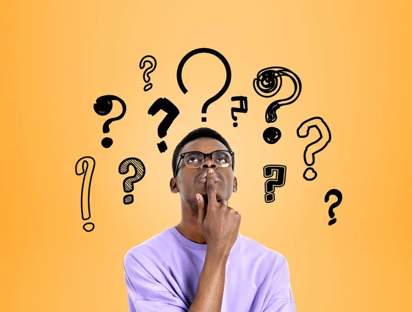 African american man with finger to mouth, serious and thoughtful look. Background of doodle question marks drawn on orange background. Concept of options