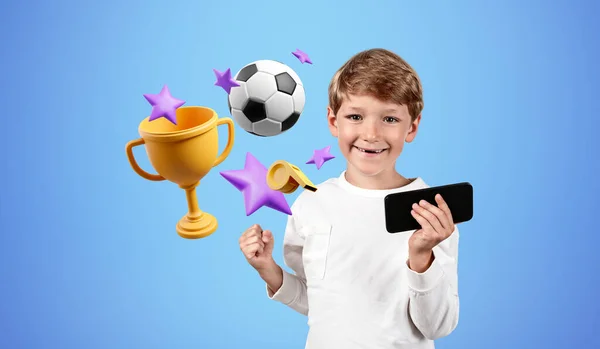 Happy child boy looking at the camera with raised fist, football and champion cup on blue background. Concept of sport, video games and mobile app