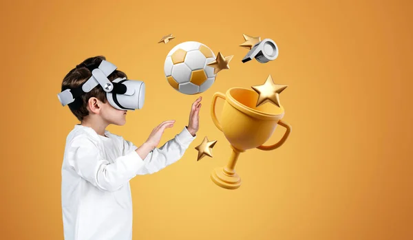 Child boy playing video games in VR headset, hands touching football, champion cup and star on orange background. Concept of simulators and VR games