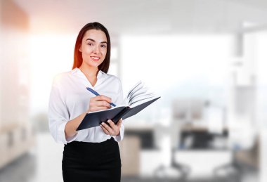 Businesswoman take note in notebook, writing and looking at camera, office room on background. Concept of education and planning