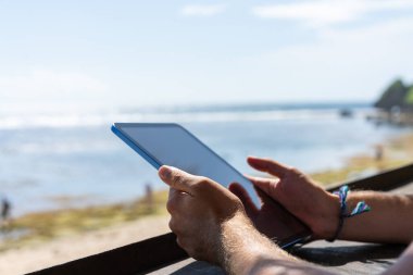 Businessman hands using tablet, blurred background of beach and sky. Online connection and internet browse. Concept of tour, traveling and holiday