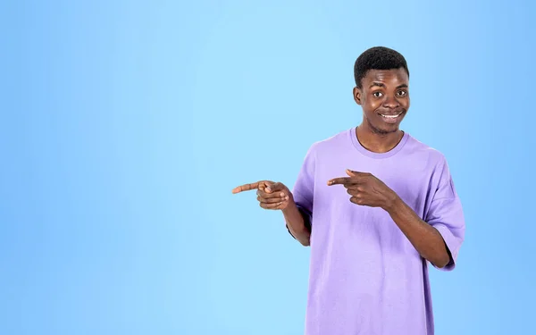 身穿紫色T恤的非洲年轻人微笑着 手指指向侧面 浅蓝色背景 推荐和商业报价的概念 复制空间 — 图库照片