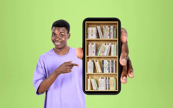 アフリカの男性は笑顔で デジタル図書館を指差すスマートフォンや指を持ち 棚の上の本 緑の背景 オンライン教育とモバイルアプリの概念 — ストック写真