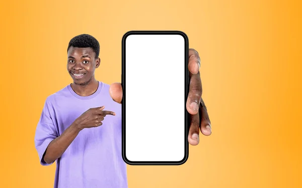 Black smiling man finger pointing at smartphone in hand, yellow background. Concept of mobile app and website. Mock up copy space display