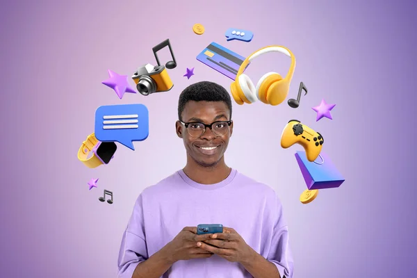 ヘッドフォン ジョイパッド ソーシャルメディアのアイコンと紫色の壁の近くに立ってスマートフォンを使用して陽気な若いアフリカ系アメリカ人の男性の肖像画 オンラインエンターテイメントの概念 — ストック写真