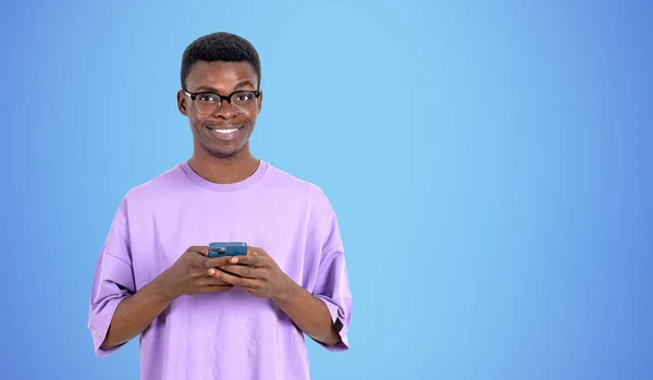 青い壁の近くにスマートフォンを保持眼鏡で陽気な若いアフリカ系アメリカ人の男性の肖像画 コミュニケーションの概念 モックアップ — ストック写真