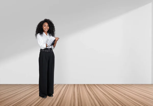African businesswoman writing notes in business notebook, looking at the camera and smiling, full length on hardwood floor. Copy space empty white wall.