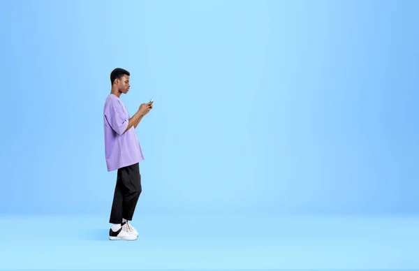 アフリカ系アメリカ人の男性のスマートフォンで 集中的に見て立っている ライトブルーの背景をブラウズしてテキストを送信します オンラインコミュニケーションの概念 スペースのコピー — ストック写真