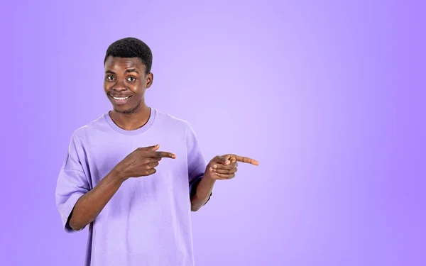 非洲年轻人穿着非常漂亮的T恤 微笑着 手指指向侧面 紫色背景 推荐和商业交易的概念 复制空间 — 图库照片