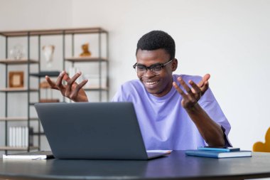 Gözlüklü duygusal Afrikalı iş adamı internette video konferansı yapıyor. Arka planda raf olan ana ofis odası. Mesafe çalışma ve buluşma kavramı