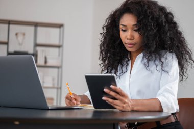 Afrikalı iş kadını not alıyor ve masadaki dijital tablet, dizüstü bilgisayar ve defterlere bakıyor. Arka planda süsleme olan ofis odası. Mesafe çalışması kavramı