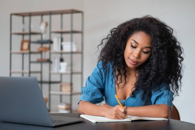 Afrikalı kadın not defterine not alıyor, dizüstü bilgisayar masasında. Arka planda süsleme olan ofis odası. E-öğrenim ve eğitim kavramı