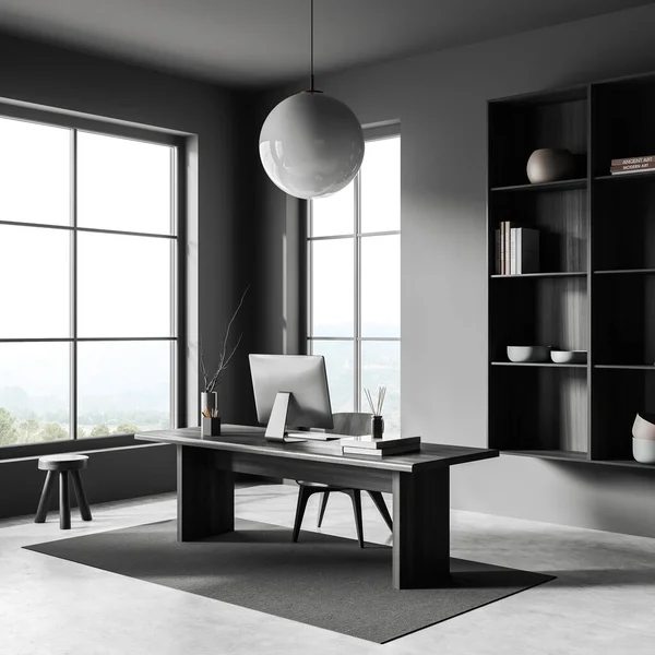 Dark workplace interior with pc computer on desk, side view, grey concrete floor. Shelf with art decoration, stool near panoramic window on countryside view. 3D rendering