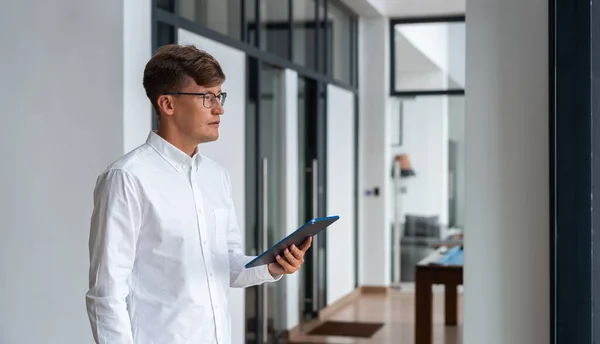 Concentrated businessman in eyeglasses, profile portrait holding a tablet standing in stylish living room apartment. Concept of online shopping and digital connection