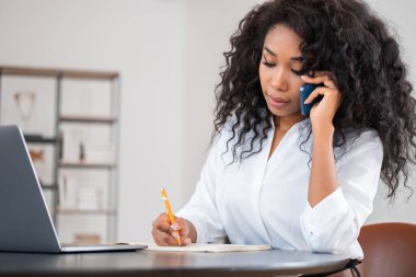 Afrikalı iş kadını not alır ve telefonda, dizüstü bilgisayarda ve masada konuşur. Arka planı bulanık raflı bir ofis odası. İletişim kavramı