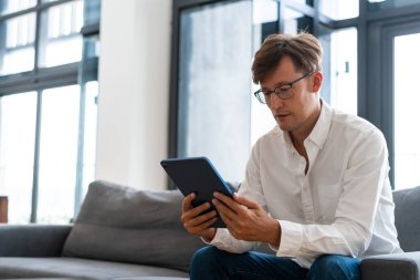 Gözlüklü genç işadamı elinde tabletle çalışıyor, pencerenin yanındaki modern apartman dairesindeki koltukta oturuyor. Çevrimiçi alışveriş ve sosyal medya kavramı