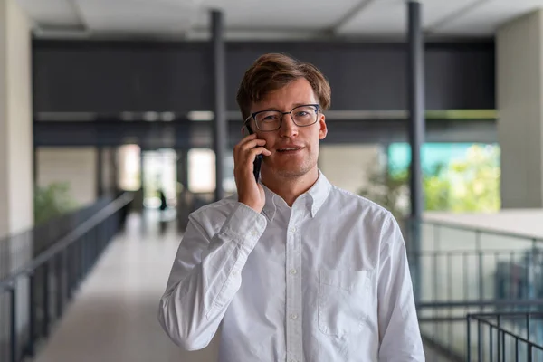 Smiling Young European Businessman Glasses Talking Smartphone Blurry Office Hall — Stock Photo, Image